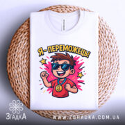 Футболка я переможець, білого кольору, складена на плетеній кошику, фон світло-сірий, принт з хлопчиком у сонцезахисних окулярах, який радісно піднімає руку, з написом 'Я - переможець!' та яскравими зірочками.