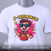 Футболка я переможець, білого кольору, розкладена на поверхні, фон світло-сірий, принт з хлопчиком у сонцезахисних окулярах, який радісно піднімає руку, з написом 'Я - переможець!' та яскравими зірочками, на комірці є етикетка.