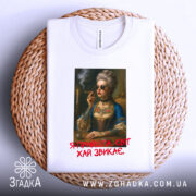 Футболка я прийшла білого кольору, зображена на плоскій поверхні, фон світло-сірий, принт з портретом жінки в окулярах, яка тримає сигарету, з текстом червоного кольору під малюнком, футболка лежить на плетеній основі.