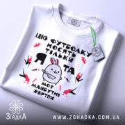 Футболка я та жертви, біла, зображена зверху, на фоні світло-сірого градієнту, принт з малюнком кролика та текстом, на футболці є етикетка з зеленим шнурком.