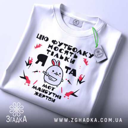 Футболка я та жертви, біла, зображена зверху, на фоні світло-сірого градієнту, принт з малюнком кролика та текстом, на футболці є етикетка з зеленим шнурком.