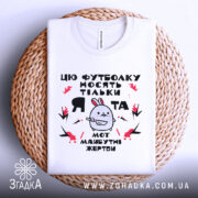 Футболка я та жертви, біла, зображена зверху на плетеному кошику, на фоні світло-сірого градієнту, принт з малюнком кролика та текстом, футболка складена акуратно.