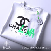 Футболка я така шалена, біла, складена на плоскій поверхні, з верхнього ракурсу, фон світло-сірий, принт чорний з логотипом та зеленим написом 'LNA', з етикеткою на комірці.