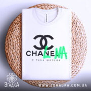 Футболка я така шалена, біла, складена на плоскій поверхні, з верхнього ракурсу, фон натуральний, плетений, принт чорний з логотипом та зеленим написом 'LNA'.