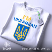 Футболка я українець англійською, біла, зображена зверху, складена на поверхні, фон світлий, принт містить текст 'I'M UKRAINIAN' жовтого та синього кольорів, а також герб України, на футболці є етикетка.