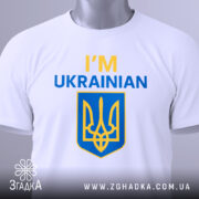 Футболка я українець англійською, біла, зображена зверху, складена на поверхні, фон світлий, принт містить текст 'I'M UKRAINIAN' жовтого та синього кольорів, а також герб України, на футболці є етикетка.