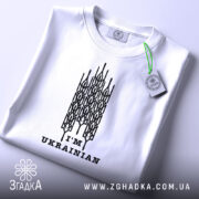 Футболка я українець, білого кольору, складена на поверхні, зображена з верхнього ракурсу, фон світло-сірий, принт чорного кольору з написом 'I'M UKRAINIAN' та етикеткою з зеленим шнурком.