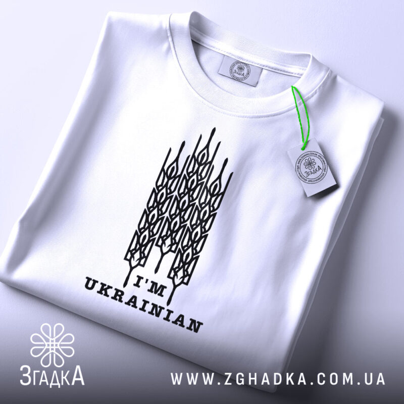 Футболка я українець, білого кольору, складена на поверхні, зображена з верхнього ракурсу, фон світло-сірий, принт чорного кольору з написом 'I'M UKRAINIAN' та етикеткою з зеленим шнурком.