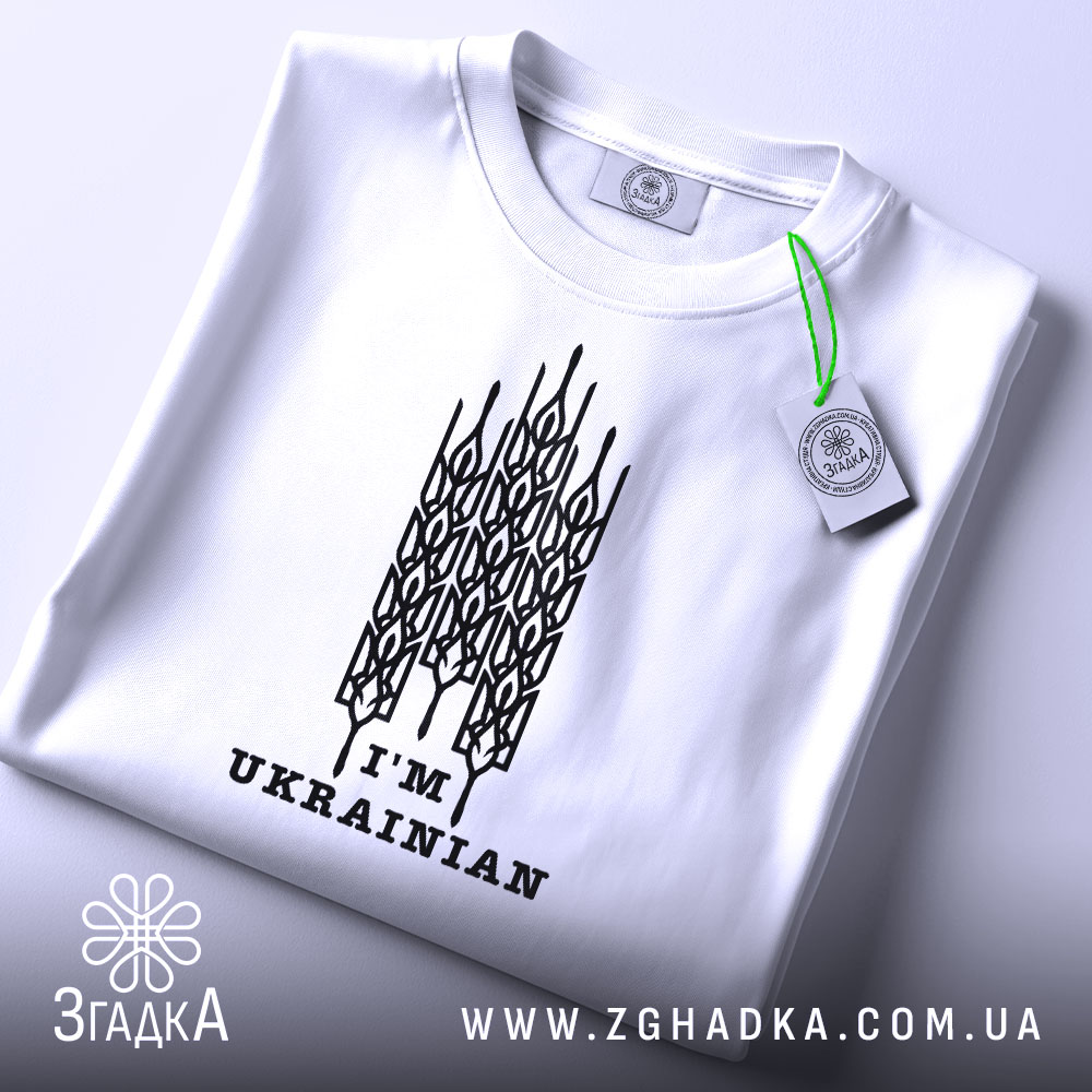 Футболка я українець, білого кольору, складена на поверхні, зображена з верхнього ракурсу, фон світло-сірий, принт чорного кольору з написом 'I'M UKRAINIAN' та етикеткою з зеленим шнурком.