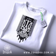 Футболка я українець принт білого кольору, складена на столі, фон світло-сірий, принт чорного кольору з тризубом та українським орнаментом, на комірці видно етикетку.