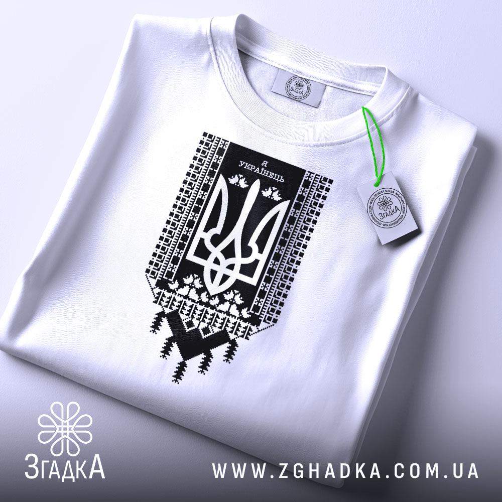 Футболка я українець принт білого кольору, складена на столі, фон світло-сірий, принт чорного кольору з тризубом та українським орнаментом, на комірці видно етикетку.