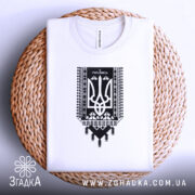 Футболка я українець принт білого кольору, складена на плетеній кошику, фон світлий, принт чорного кольору з тризубом та українським орнаментом, видно етикетку на комірці.