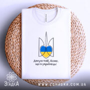 Футболка я українець, біла, зображена зверху на плетеній основі, фон білий, принт у вигляді тризуба з синьо-жовтими елементами та написом 'Дякую тобі, Боже, що я українець!'.