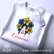 Футболка я українка, білого кольору, складена на поверхні, фон світло-сірий, принт з дівчиною з квітами в волоссі, текст "Я українка!" червоним кольором, з етикеткою на комірці.