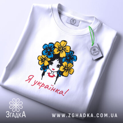 Футболка я українка, білого кольору, складена на поверхні, фон світло-сірий, принт з дівчиною з квітами в волоссі, текст "Я українка!" червоним кольором, з етикеткою на комірці.
