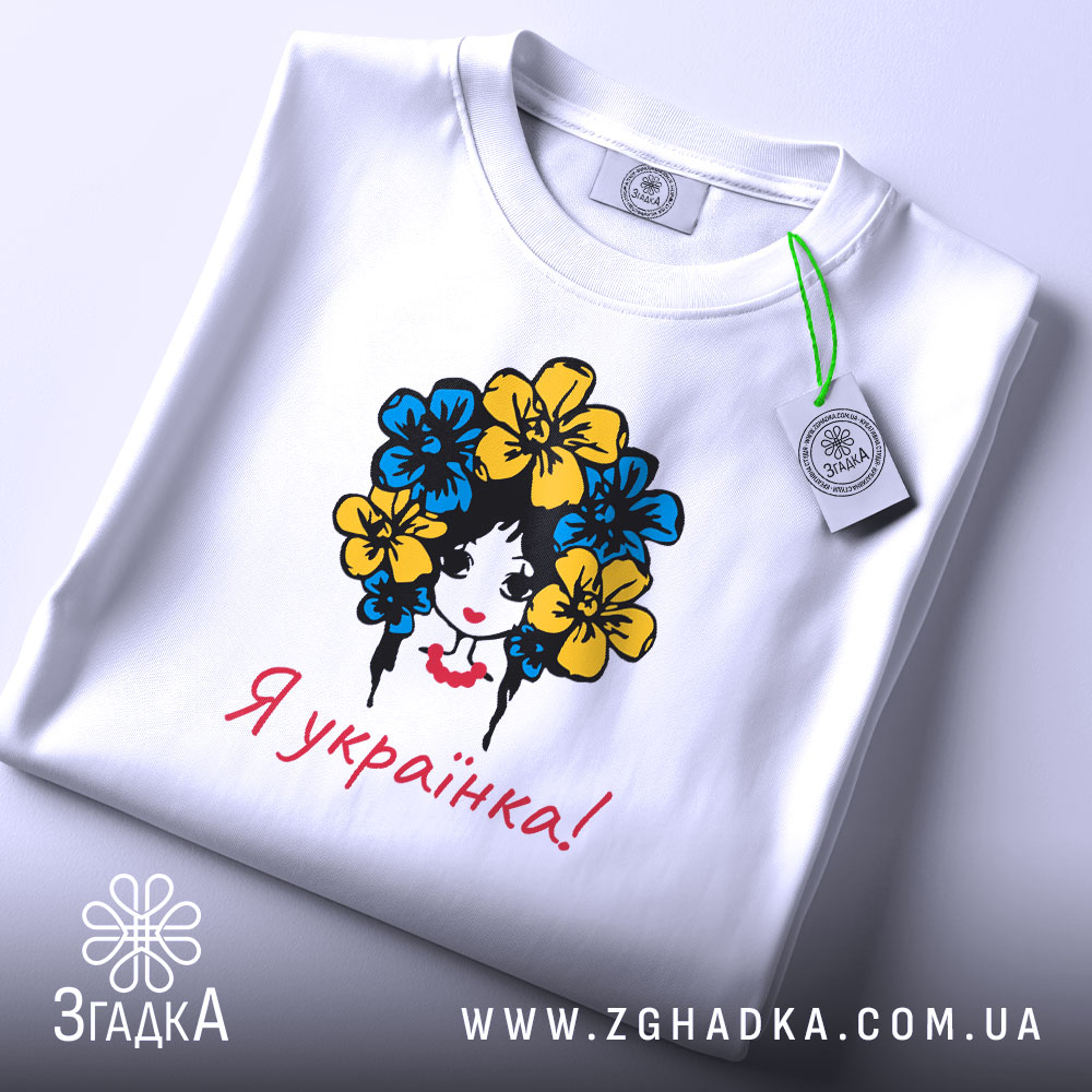Футболка я українка, білого кольору, складена на поверхні, фон світло-сірий, принт з дівчиною з квітами в волоссі, текст "Я українка!" червоним кольором, з етикеткою на комірці.