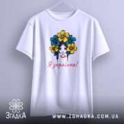 Футболка я українка, білого кольору, підвішена на вішалці, фон світло-сірий, принт з дівчиною з квітами в волоссі, текст "Я українка!" червоним кольором.