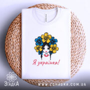 Футболка я українка, білого кольору, складена на плетеній кошику, фон білий, принт з дівчиною з квітами в волоссі, текст "Я українка!" червоним кольором.