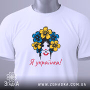 Футболка я українка, білого кольору, складена на поверхні, фон світло-сірий, принт з дівчиною з квітами в волоссі, текст "Я українка!" червоним кольором, з етикеткою на комірці.