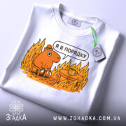 Футболка я в порядку. Біла футболка, складена на столі, з принтом, що зображує коричневу тварину на стільці біля столу з чашкою, на фоні полум'я. Принт яскравий, з жовтими та оранжевими відтінками, з етикеткою на зеленому шнурку.