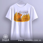 Футболка я в порядку. Біла футболка, підвішена на вішалці, з принтом, що зображує коричневу тварину на стільці біля столу з чашкою, на фоні полум'я. Принт яскравий, з жовтими та оранжевими відтінками.