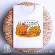 Футболка я в порядку. Біла футболка, складена на плетеній основі, з принтом, що зображує коричневу тварину на стільці біля столу з чашкою, на фоні полум'я. Принт яскравий, з жовтими та оранжевими відтінками.