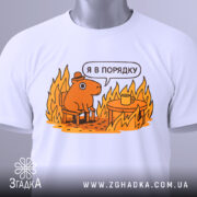 Футболка я в порядку. Біла футболка, складена на столі, з принтом, що зображує коричневу тварину на стільці біля столу з чашкою, на фоні полум'я. Принт яскравий, з жовтими та оранжевими відтінками, з етикеткою на зеленому шнурку.