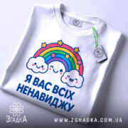 Футболка я вас ненавиджу, біла, зображена на столі, фон світло-сірий, принт з веселкою та хмарами, на комірці є етикетка.