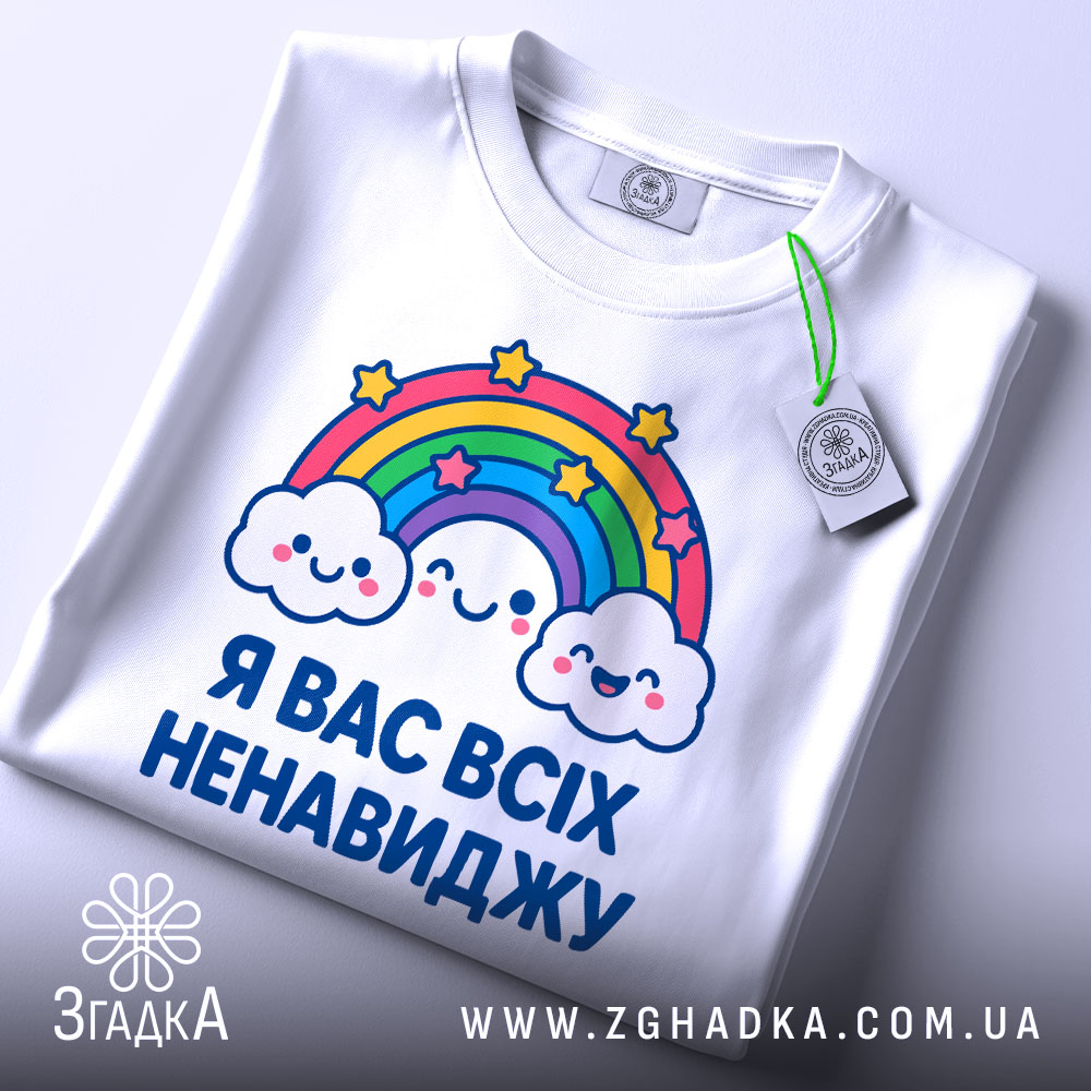 Футболка я вас ненавиджу, біла, зображена на столі, фон світло-сірий, принт з веселкою та хмарами, на комірці є етикетка.