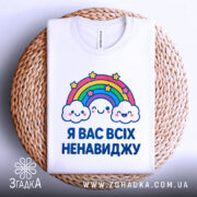 Футболка я вас ненавиджу, біла, зображена на столі, фон світло-сірий, принт з веселкою та хмарами, розташована на плетеному кошику.