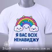 Футболка я вас ненавиджу, біла, зображена на столі, фон світло-сірий, принт з веселкою та хмарами, на комірці є етикетка.