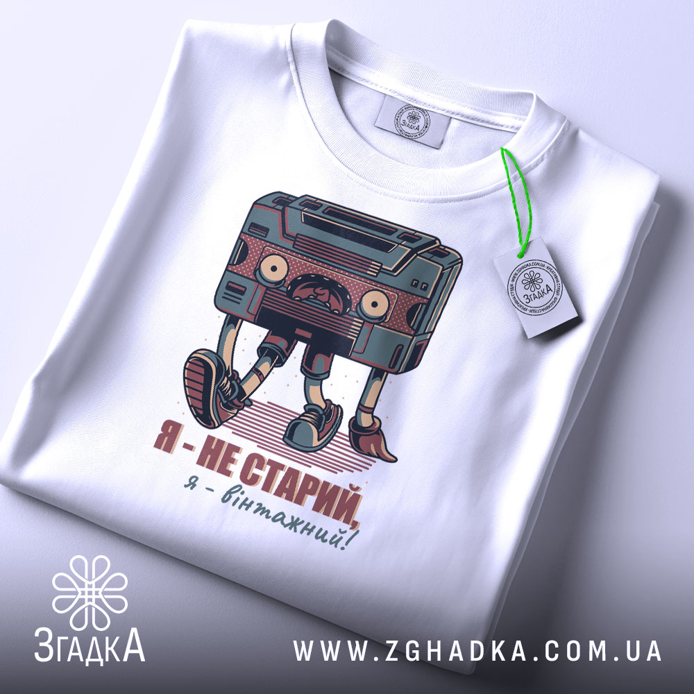 Футболка я вінтажний, білого кольору, складена на поверхні, фон світло-сірий, принт з касетою та написом "Я - не старий, я - вінтажний!" у кольорах червоного та зеленого, на комірці видна етикетка.