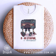 Футболка я вінтажний, білого кольору, складена на плетеній основі, фон світло-сірий, принт з касетою та написом "Я - не старий, я - вінтажний!" у кольорах червоного та зеленого.