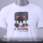 Футболка я вінтажний, білого кольору, складена на поверхні, фон світло-сірий, принт з касетою та написом "Я - не старий, я - вінтажний!" у кольорах червоного та зеленого, на комірці видна етикетка.