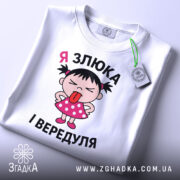 Футболка я злюка вередуля. Біла футболка, складена на поверхні, з принтом дівчинки в рожевій сукні, яка показує язика. На футболці видно етикетку з зеленим шнурком, фон світло-сірий.