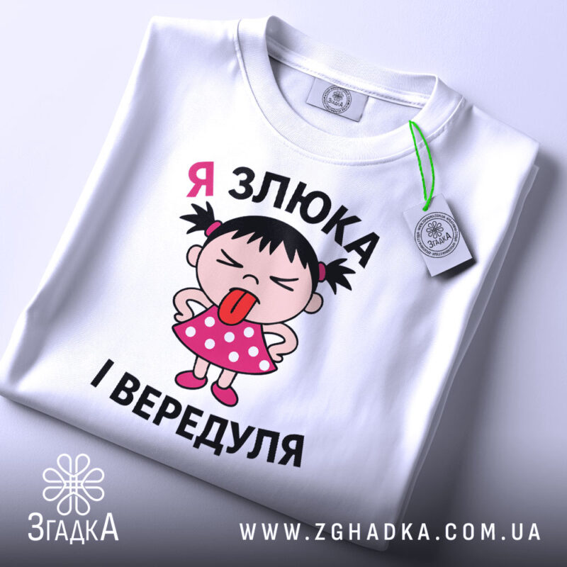 Футболка я злюка вередуля. Біла футболка, складена на поверхні, з принтом дівчинки в рожевій сукні, яка показує язика. На футболці видно етикетку з зеленим шнурком, фон світло-сірий.