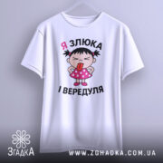 Футболка я злюка вередуля. Біла футболка з коротким рукавом, підвішена на вішалці, з принтом дівчинки в рожевій сукні, яка показує язика. Фон світло-сірий.