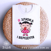 Футболка я злюка вередуля. Біла футболка, складена на плетеній кошику, з принтом дівчинки в рожевій сукні, яка показує язика. Фон білий.