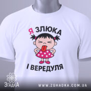 Футболка я злюка вередуля. Біла футболка, складена на поверхні, з принтом дівчинки в рожевій сукні, яка показує язика. На футболці видно етикетку з зеленим шнурком, фон світло-сірий.