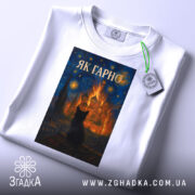 Футболка як гарно білого кольору, складена на поверхні, з принтом, що зображає чорного кота на фоні яскравого пейзажу, знімок зроблений зверху, на футболці видно етикетку.