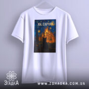 Футболка як гарно білого кольору, підвішена на вішалці, з принтом, що зображає чорного кота на фоні яскравого пейзажу, знімок зроблений спереду.