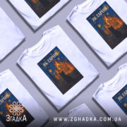 Футболка як гарно білого кольору, з принтом на грудях, що зображає чорного кота на фоні яскравого пейзажу з церквою та зірками, знімок зроблений спереду на манекені.