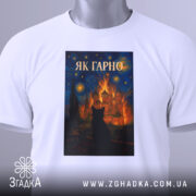 Футболка як гарно білого кольору, складена на поверхні, з принтом, що зображає чорного кота на фоні яскравого пейзажу, знімок зроблений зверху, на футболці видно етикетку.
