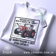 Футболка як танкіст тракторист, біла, зображена з верхнього ракурсу, фон світлий, принт з червоним трактором та текстом, з етикеткою зеленого кольору.