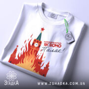 Футболка як воно палає, біла, складена на поверхні, фон світло-сірий, принт з червоними і жовтими вогнями, зображення Кремля з годинником і зіркою, на футболці є етикетка з зеленим шнурком.