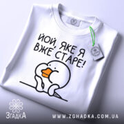 Футболка яке я старе, білого кольору, зображена зверху на плоскій поверхні, фон світло-сірий, принт з малюнком пташки з оранжевим дзьобом і текстом 'Йой, яке я вже старе!', на комірці є етикетка.