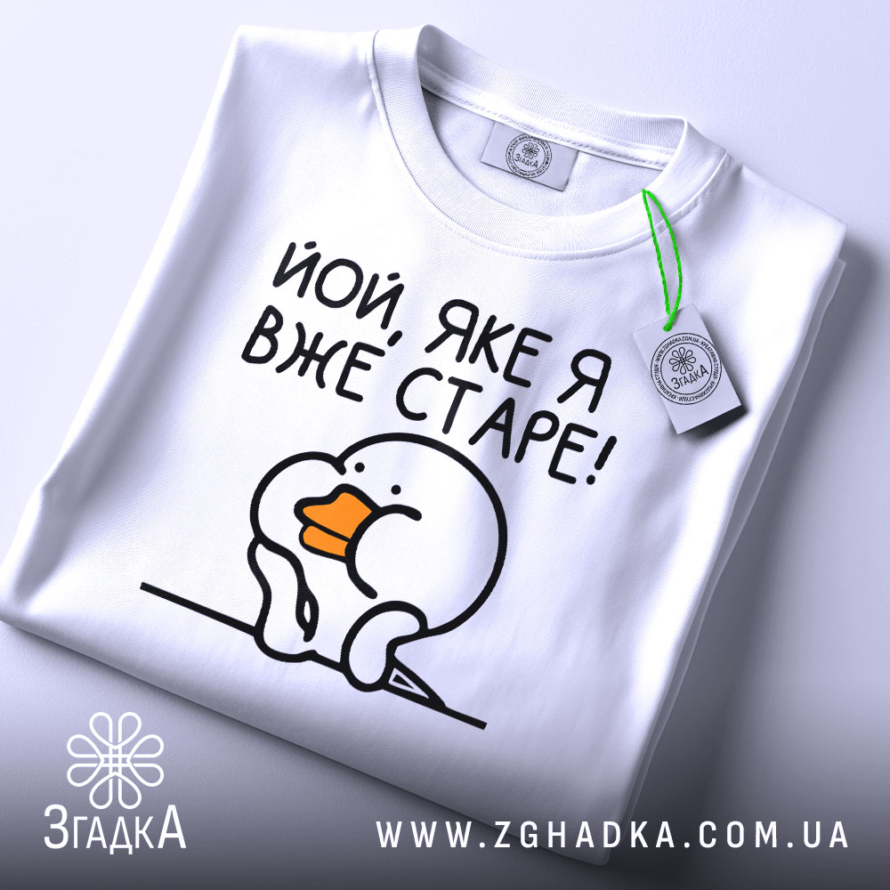 Футболка яке я старе, білого кольору, зображена зверху на плоскій поверхні, фон світло-сірий, принт з малюнком пташки з оранжевим дзьобом і текстом 'Йой, яке я вже старе!', на комірці є етикетка.