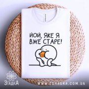 Футболка яке я старе, білого кольору, зображена зверху на плоскій поверхні, фон світло-сірий, принт з малюнком пташки з оранжевим дзьобом і текстом 'Йой, яке я вже старе!', футболка розташована на плетеній основі.