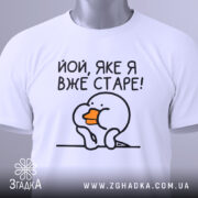 Футболка яке я старе, білого кольору, зображена зверху на плоскій поверхні, фон світло-сірий, принт з малюнком пташки з оранжевим дзьобом і текстом 'Йой, яке я вже старе!', на комірці є етикетка.