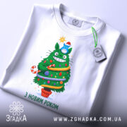 Футболка ялинка новий рік, біла, зображена на столі, фон світлий, принт з зеленим деревом, яке має червону шапочку, прикрашене іграшками та написом 'З НОВИМ РОКОМ', на футболці є бірка.